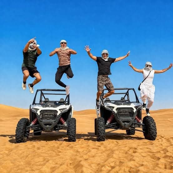 Four friends jumping for a photo in front of two off-road buggies on the dunes