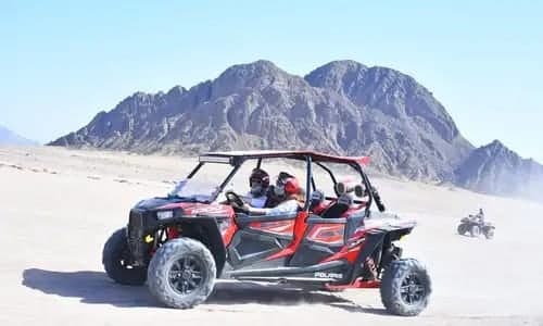 Red four-seat buggy with riders in helmets on pale desert sand below rocky mountains