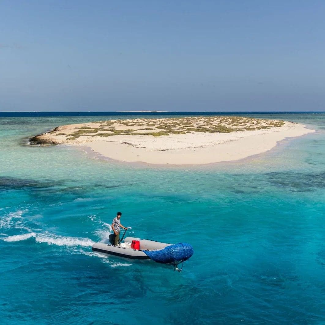 Small sandy island ringed by turquoise water, with a dinghy speeding past on a boat trip