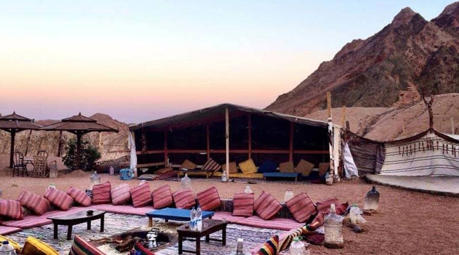 Bedouin camp with floor cushions, low tables and tents below desert mountains at sunset