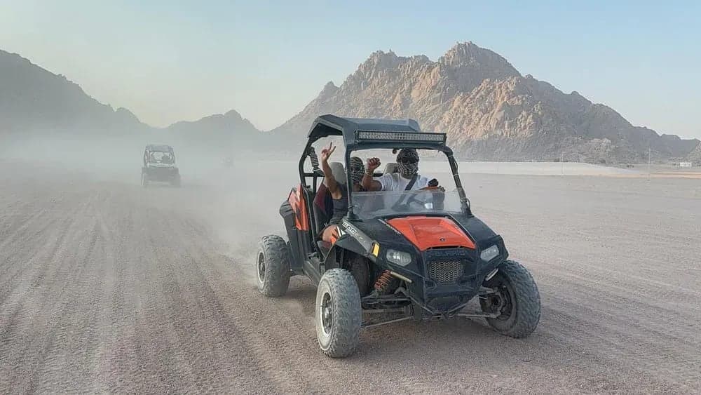 Private buggy safari: two riders waving from an orange two-seat buggy driving through desert dust below rocky mountains